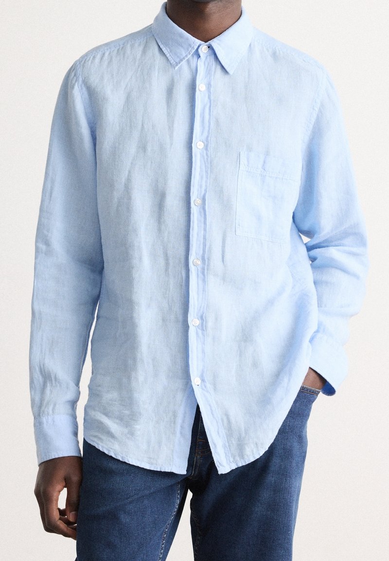Camicia button-up in lino azzurro chiaro con colletto, taschino sul petto, e maniche lunghe; abbinata a jeans di jeans scuri.