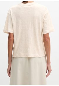 Personne portant un t-shirt beige ample à manches courtes et une jupe de couleur claire, vue de dos sur un fond uni.