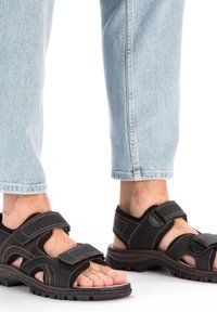 Sandales noires à bout ouvert avec des sangles réglables en Velcro, semelle en caoutchouc épaisse et coutures contrastées. Portées avec un jean léger à revers bleu.