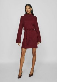 Robe en tricot bourgogne avec un col haut, des manches longues et une taille ceinturée. Présente des poignets côtelés et un ourlet légèrement effiloché. Chaussures à talons marron.