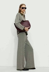 Donna che indossa una blusa a quadri bianco e nero e pantaloni a gamba larga, che tiene una grande borsa intrecciata e indossa occhiali da sole neri.