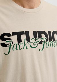 Kremowa koszulka T-shirt z czarnym, wyraźnym napisem "STUDIO" oraz zielonym, kursywą napisem "Jack & Jones", wykonana z miękkiej tkaniny bawełnianej z okrągłym dekoltem.
