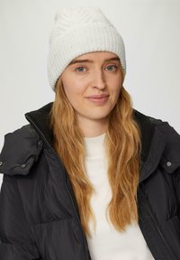 Weißer, gerippter Strickbeanie mit strukturiertem Design. Modell trägt eine schwarze Steppjacke mit Reißverschluss- und Knopfdetails über einem weißen Oberteil.