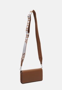 Brązowa skórzana torebka o prostokątnym kształcie, z teksturowaną powierzchnią. Regulowany pasek z białym logo. Zamek błyskawiczny jako detal zamknięcia.