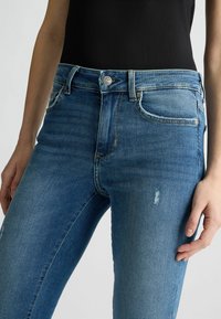 Blaue Jeans aus Denim mit einer taillierten Passform, ausgestattet mit einer verblassten Waschung, distressierten Details und einem klassischen Knopfverschluss. Nahaufnahme von Taille und Beinen.