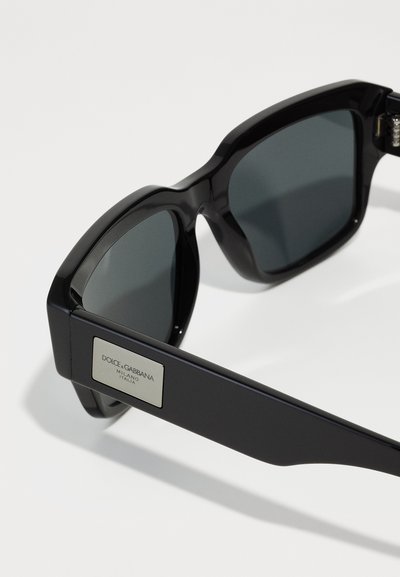 Black sunglasses with a chunky frame, grey lenses, and a rectangular shape. Features a silver logo plaque on the arm.