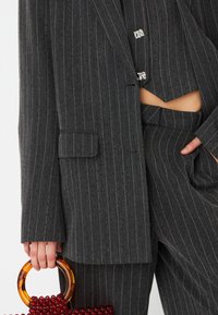 Completo grigio scuro con righe sottili, giacca corta e pantaloni coordinati; presenta bottoni argentati lucidi e una borsa a tracolla bordeaux decorata con perline, dotata di un manico ambra.