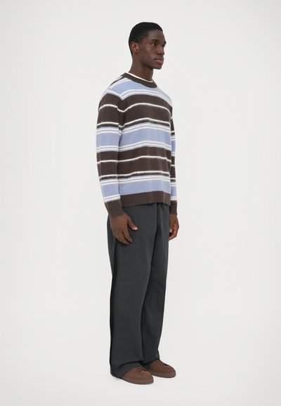 Pull en maille rayé de couleur marron, bleu clair et blanc, avec un col rond et des poignets côtelés, associé à un pantalon ample de couleur foncée et des chaussures marron.