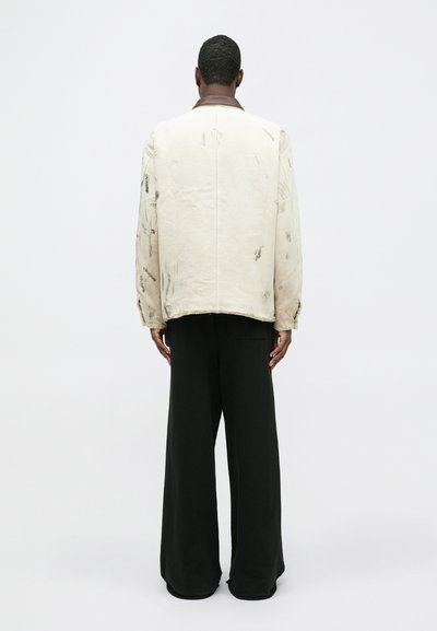 Homme de dos portant une veste crème avec des taches grises et un pantalon noir à jambes larges, contre un fond uni.