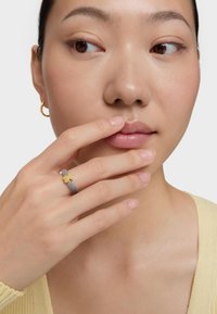 Anillo de malla plateada con acento floral dorado en un dedo, usado por una persona con un suéter de tono neutro, piel suavemente iluminada y maquillaje mínimo.