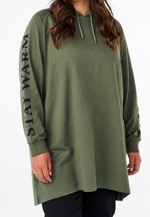 Nainen, jolla on yllään valtavan kokoinen oliivinvihreä huppari, jossa on vasemmassa hihassa teksti "STAY WARM", yhdistettynä mustiin housuihin.