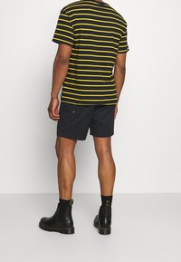 Černožluté pruhované tričko ve spojení s černými šortkami. Model má na nohou černé chelsea boty s texturovanou podrážkou. Zobrazen pohled zezadu.