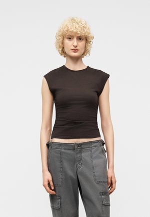 Osoba o blond kręconych włosach, ubrana w dopasowany czarny top bez rękawów i szare spodnie cargo, stojąca na tle jasnego, jednolitego tła.