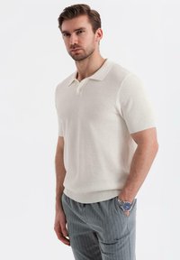 Camisa polo de manga corta color crema con un diseño de punto texturizado, presenta un cuello clásico y un dobladillo de canalé, combinada con pantalones rayados grises.