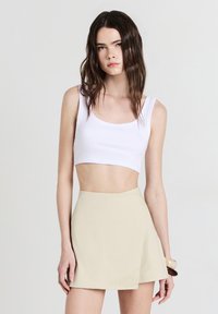 Giovane donna con lunghi capelli scuri che indossa un top corto bianco e una gonna mini asimmetrica beige su uno sfondo neutro.