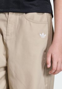 Beżowe spodnie wykonane z trwałej tkaniny, z delikatnie teksturowaną powierzchnią, bocznymi kieszeniami oraz małym białym logo Adidas na lewej kieszeni.