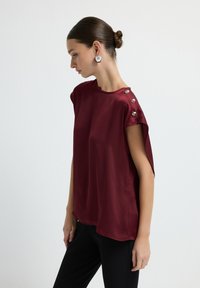 Top corto bordeaux a maniche corte con una superficie liscia, dotato di bottoni decorativi lungo la cucitura della spalla e un design morbido.