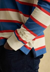 Henkilö, joka pukee päälleen pitkähihainen rugby-paita, jossa on leveät siniset, punaiset ja kerma-valkoiset vaakaraitat sekä olkapäissä brodeerattu teksti "JOULES MCMLXXX".