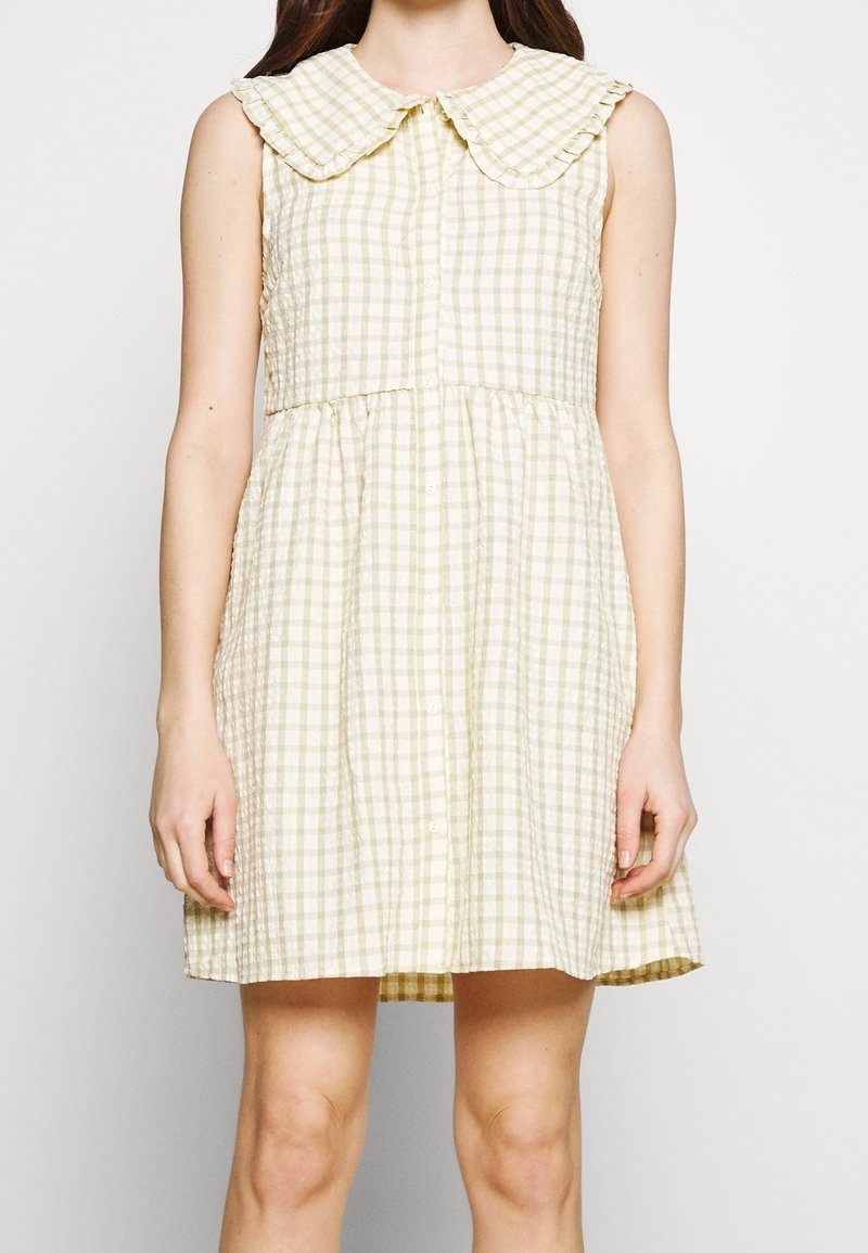 Robe sans manches à carreaux beige et blanc avec un grand col arrondi et des boutons sur le devant, portée par une personne debout devant un fond uni.
