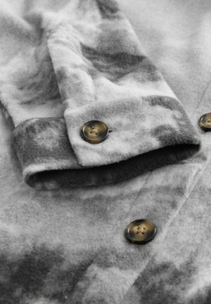 Harmaan fleece-kangas, jossa on laikukas kuvio, napin suljento ja taitettu hihansuu. Pehmeä koostumus ja hillitty väriasteikko.