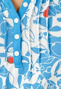 Top con patrón floral azul y blanco de textura acanalada, con una tapeta de botones azul y tres botones blancos. Se puede ver un pequeño acento rojo.