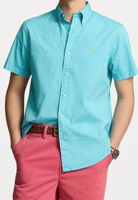 Kortärmad skjorta i ljus turkos bomull med button-down krage, prydd med vita knappar och en liten gul logotyp på bröstet. Matchad med rosa shorts.