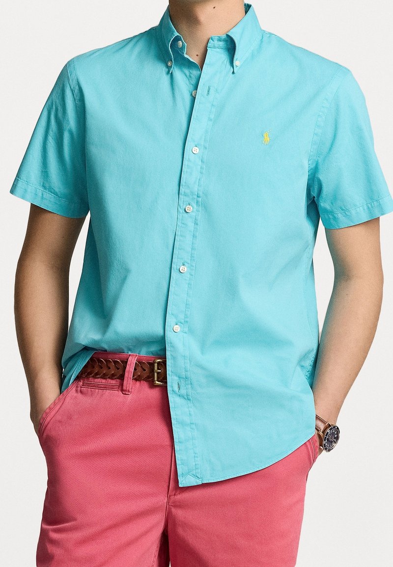 Kortärmad skjorta i ljus turkos bomull med button-down krage, prydd med vita knappar och en liten gul logotyp på bröstet. Matchad med rosa shorts.