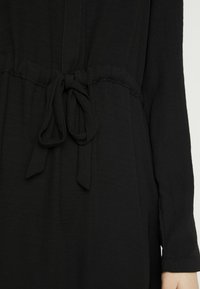 Schwarzes Kleid mit langen Ärmeln, einem Kordelzug in der Taille und einem weichen, strukturierten Stoff. Einfaches Design ohne sichtbare Muster oder Hardware.