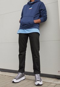 Felpa con cappuccio blu navy con tasca frontale, indossata sopra una camicia azzurro chiaro. Pantaloni neri affusolati e sneakers grigie completano il look.
