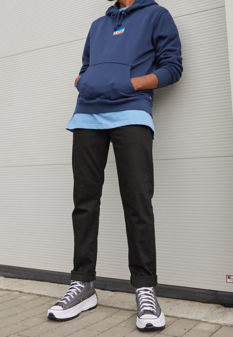 Felpa con cappuccio blu navy con tasca frontale, indossata sopra una camicia azzurro chiaro. Pantaloni neri affusolati e sneakers grigie completano il look.