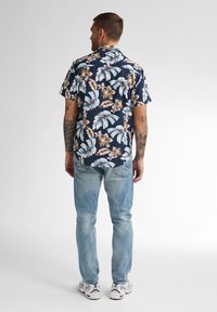 Camisetta a maniche corte blu navy con un motivo floreale che presenta foglie azzurro chiaro e fiori beige, abbinata a jeans azzurro chiaro e sneakers bianche.