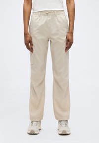 Άτομο που φοράει μπεζ παντελόνι cargo με ελαστική μέση και μπεζ αθλητικά παπούτσια, στέκεται μπροστά σε απλό φωτεινό φόντο.