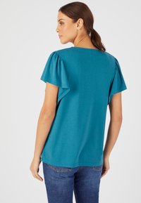 Top corto de color verde azulado con mangas abullonadas, tejido suave, corte relajado y escote redondeado, combinado con jeans azules. Vista trasera.