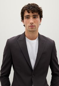 Blazer grigio scuro con revers a raggio, chiusura a un bottone e taschino sul petto, indossato sopra una t-shirt bianca, texture liscia.