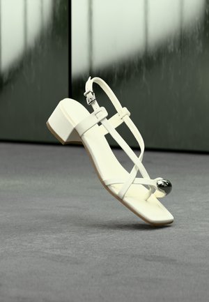 Λευκό σανδάλι με λουράκια, χαμηλό χονδρό τακούνι και ασημένια διακοσμητική μεταλλική σφαίρα στο λουρί του δακτύλου, πάνω σε γκρι επιφάνεια με θολό πράσινο φόντο.