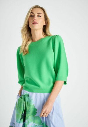 Blonde vrouw met een felgroene trui met opgeblazen mouwen en een lichtblauwe rok met groene bloemenprint, staand tegen een witte achtergrond.