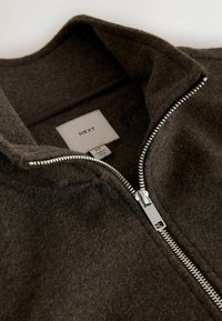 Hnedá zipsová bunda vyrobená z textúrované vlny s vysokým golierom, so strieborným zipsom a štítkom s nápisom „NEXT“ a údajmi o veľkosti.