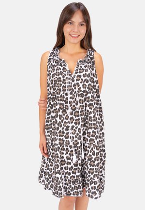 Junge Frau mit braunen Haaren, die ein ärmelloses Kleid mit Leopardenmuster, einem gerüschten Ausschnitt und vorne mit Quastenbändern trägt, lächelt in die Kamera.
