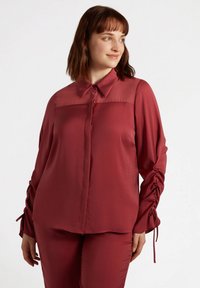 Blusa lunga a maniche con colletto di colore bordeaux, chiusura con bottoni sul davanti e dettagli arricciati sulle maniche. Tessuto liscio con un pannello superiore trasparente.