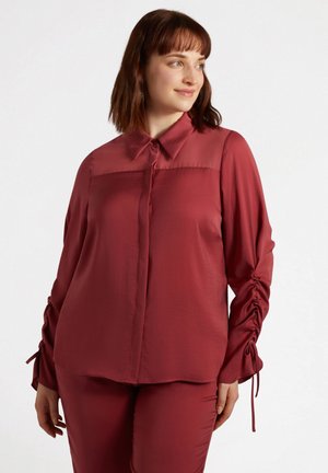Blouse bordeaux à manches longues avec un col, une fermeture à boutons sur le devant et des détails froncés sur les manches. Tissu lisse avec un panneau supérieur transparent.