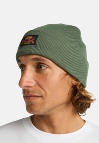 Beanie verde a coste con un knit testurizzato, etichetta nera con logo arancione. Si adatta perfettamente alla testa, senza ulteriori decorazioni.