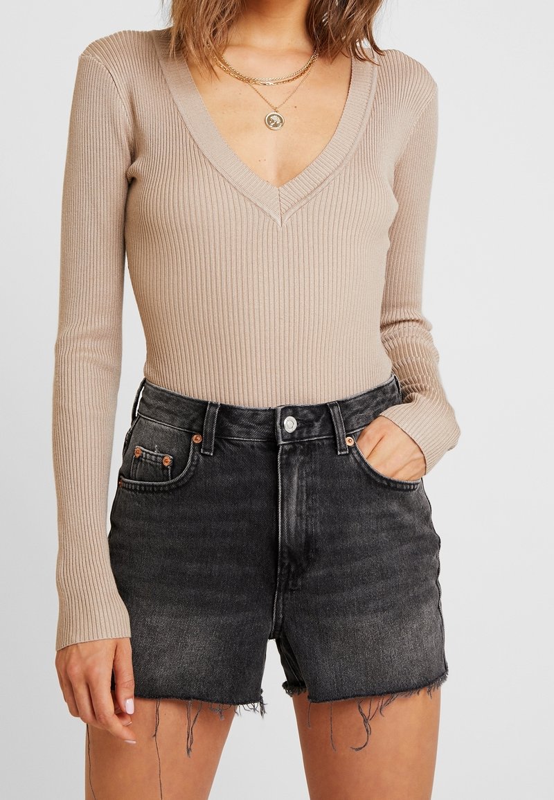 Femme portant un haut à manches longues beige à col en V côtelé et un short en denim noir délavé taille haute avec des bords effilochés, la main dans la poche.