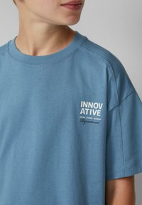 Jasnoniebieski bawełniany t-shirt z okrągłym dekoltem, krótkimi rękawami i białym nadrukiem tekstu po lewej stronie. Miękka tekstura, luźny krój.
