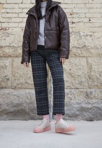 Brun vadderad jacka, grå turtleneck, svarta rutiga byxor och beige sneakers med rosa sulor. Väggtexturen är grov och i ljusa färger.