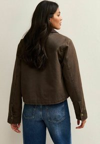 Bruine cropped jas met lange mouwen, knoopmanchetten en een gladde textuur. Draag over blauwe jeans, gezien van achteren.