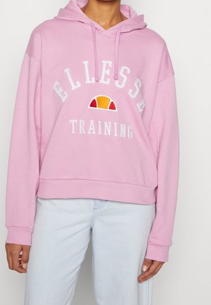 Osoba nosząca jasnoróżową bluzę z kapturem z napisem i logo "ELLESSE TRAINING", połączoną z jasnoniebieskimi dżinsami, stojąca na białym tle.