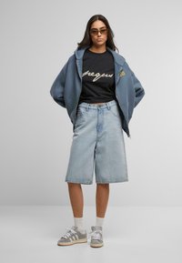 Overdimensjonert blå hettegenser, svart skjorte med skripttekst, lyseblå denimshorts og grå joggesko med hvite detaljer. Minimalistisk design.