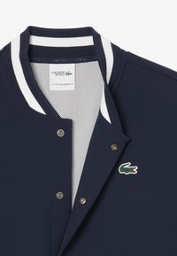 Chaqueta navy con cuello de rayas blancas y botones a presión. Presenta un forro de malla y un logo de cocodrilo verde en el lado izquierdo.
