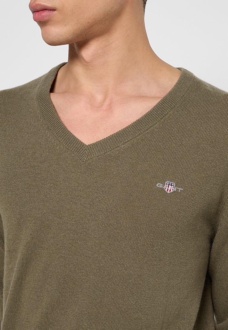 Olivgrön stickad v-ringad tröja med ribbad halsringning, med en liten broderad logotyp på bröstet. Slät textur och figursydd design.