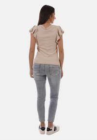 Femme debout, vue de dos, vêtue d'un haut beige à manches flottantes, d'un jean skinny gris clair et de baskets blanches avec des accents noirs.